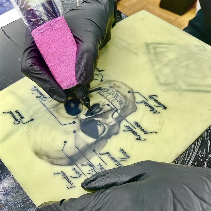 Hände in Handschuhen führen die Tattoo-Maschine über Kunsthaut